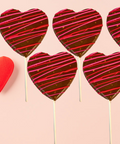 Chocolate heart lollipops with red and pink drizzle, set of 5, on a pink background with a red heart decoration.