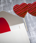 Heart-shaped chocolate lollipops with red and pink drizzle next to an envelope with a red heart card on a gray background.