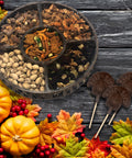 Thanksgiving nut tray with gourmet snacks and chocolate turkey lollipops, surrounded by autumn leaves and pumpkins.